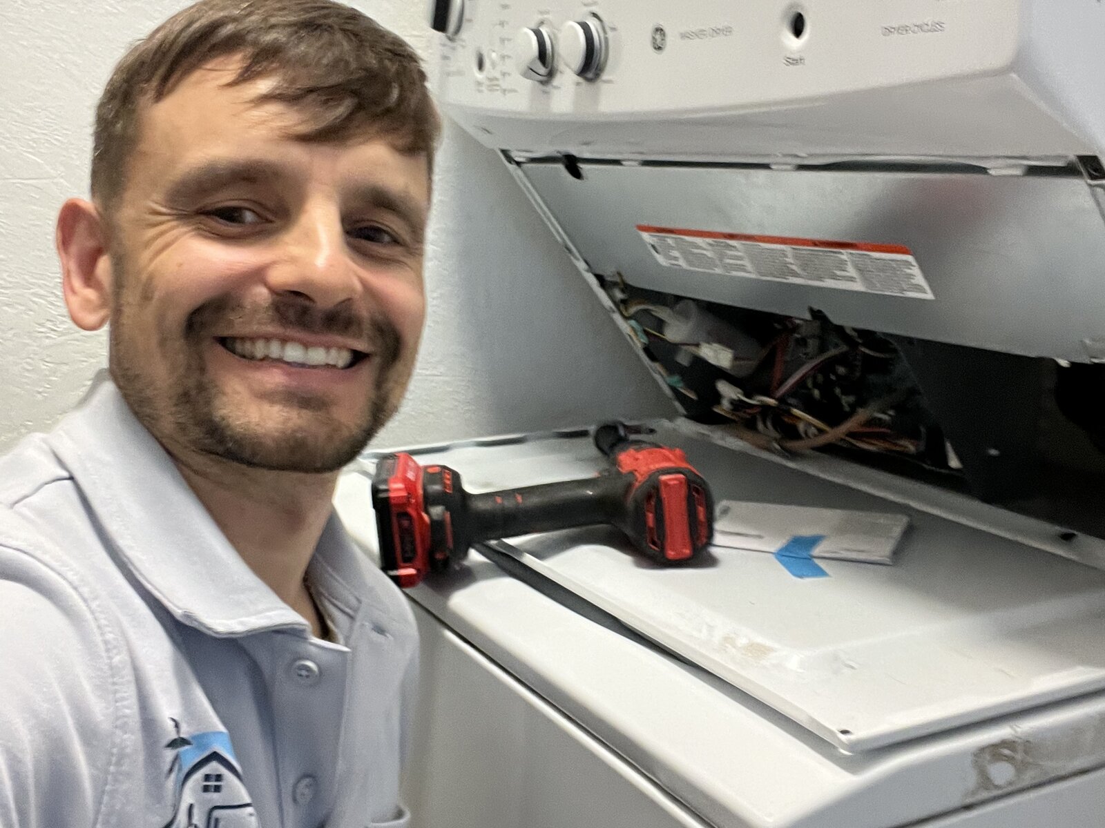 Konstantin in branded polo at a stacked washer/dryer laundry center, panel off mid-repair